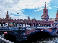 Oberbaumbrücke  The Oberbaum Bridge (German: Oberbaumbrücke) is a double-deck bridge crossing Berlin's River Spree, considered one of the city's landmarks. It links Friedrichshain and Kreuzberg, former boroughs that were divided by the Berlin Wall, and has become an important symbol of Berlin’s unity. By 1879 the wooden bridge had been modified greatly. At 154 meters it was Berlin's longest, but was no longer adequate to the amount of traffic crossing it. Plans began to be drawn up for a new stone construction. The Siemens & Halske company, which was planning to build the Berlin U-Bahn (subway), insisted on a combined crossing for road vehicles, pedestrians, and the new rail line. The new bridge opened in 1896 after two years of construction, in time for the Berlin Trades Exhibition. The architect and government official Otto Stahn (1859-1930) designed it in North German Brick Gothic, in the style of a city gate with many decorative elements, such as pointed arches, cross vaults, and coats of arms. The two towers were inspired by the Middle Gate Tower (Mitteltorturm) in the northern Brandenburg city of Prenzlau. Although purely cosmetic, they served as a reminder that the site was once Berlin’s river gateway. : 2016, Berliini, Berlin, Fujifilm, digital image, kaupunki, kesäloma, summer holiday, town