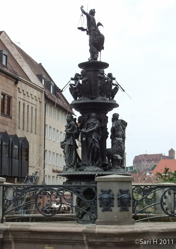 nurmberg-21.jpg - Tugendbrunnen (Fountain of the Virtues), in 1589, the iron caster, Benedikt Wurzelbauer, completed the Fountain of the Virtues. Six allegories of the three theological and the three cardinal virtues with their attributes are placed on a round platform: Faith with a cross and a chalice, Love with two children, Hope with an anchor, Courage with a lion, Moderation with a jug, and Patience with a lamb. Above the figures, cherubs carry the two coats of arms of the City of Nuremberg. The seventh virtue, Justice, stands on the top of the pillar with blindfolded eyes, a sword and a crane as a symbol of alertness.